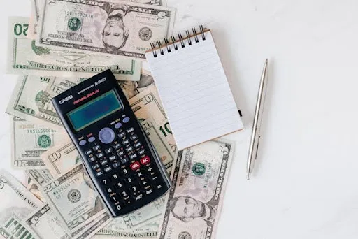 A calculator and a blank notepad resting on scattered US banknotes.
