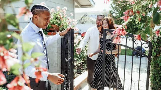 An agent holds a garden gate open as a couple arrives to view a house.