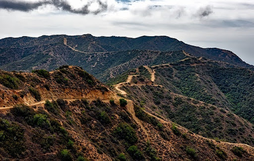 Dirt trails winding across dry chaparral hills under a cloudy sky.
